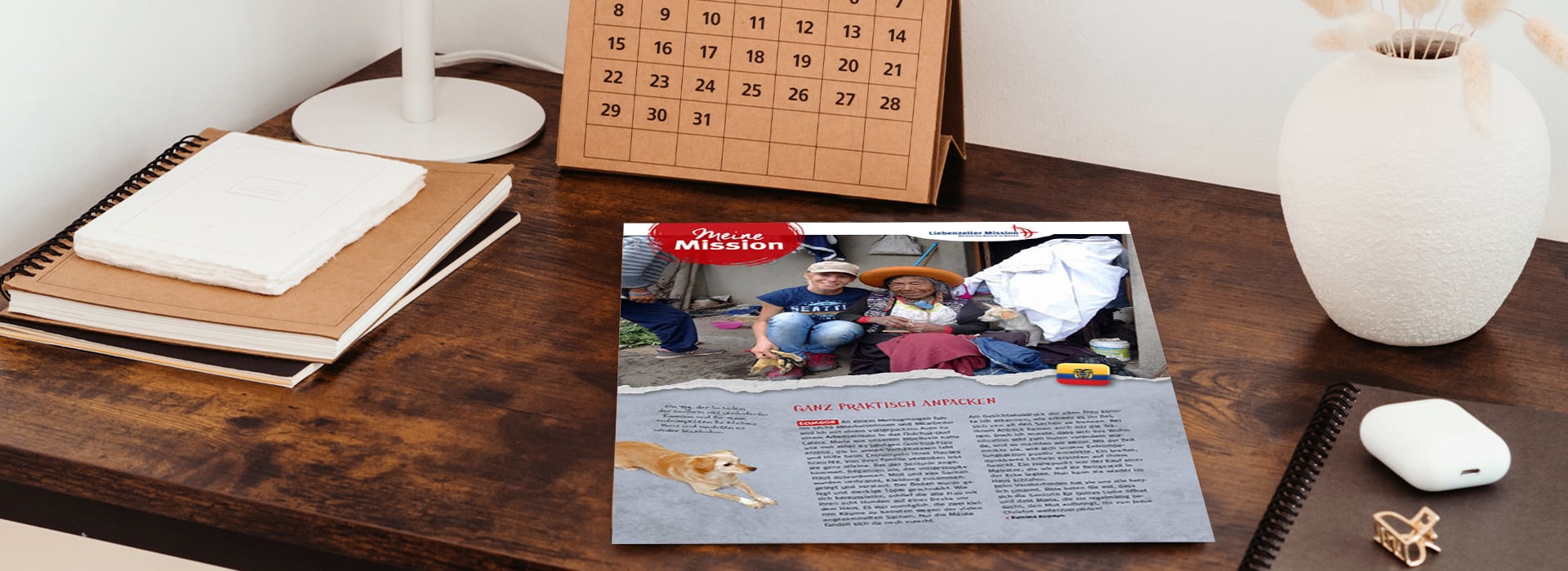 Auf einem Holztisch liegt eine Zeitschrift mit einem Foto von Menschen und einem Hund neben Notizbüchern, einem Kalender und einer Lampe.