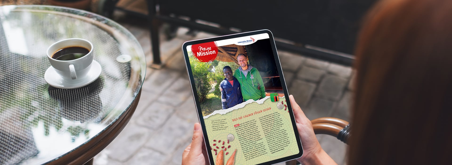 Eine Person, die ein Tablet in der Hand hält, auf dem ein Artikel mit Fotos von zwei Personen zu sehen ist; daneben steht eine Tasse Kaffee auf einem Glastisch.