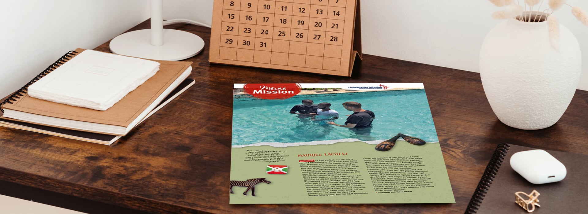 Auf einem Holztisch liegt ein Flyer, auf dem zwei Personen im Wasser zu sehen sind; daneben befinden sich Notizbücher, ein Kalender, eine Vase und eine Maus.