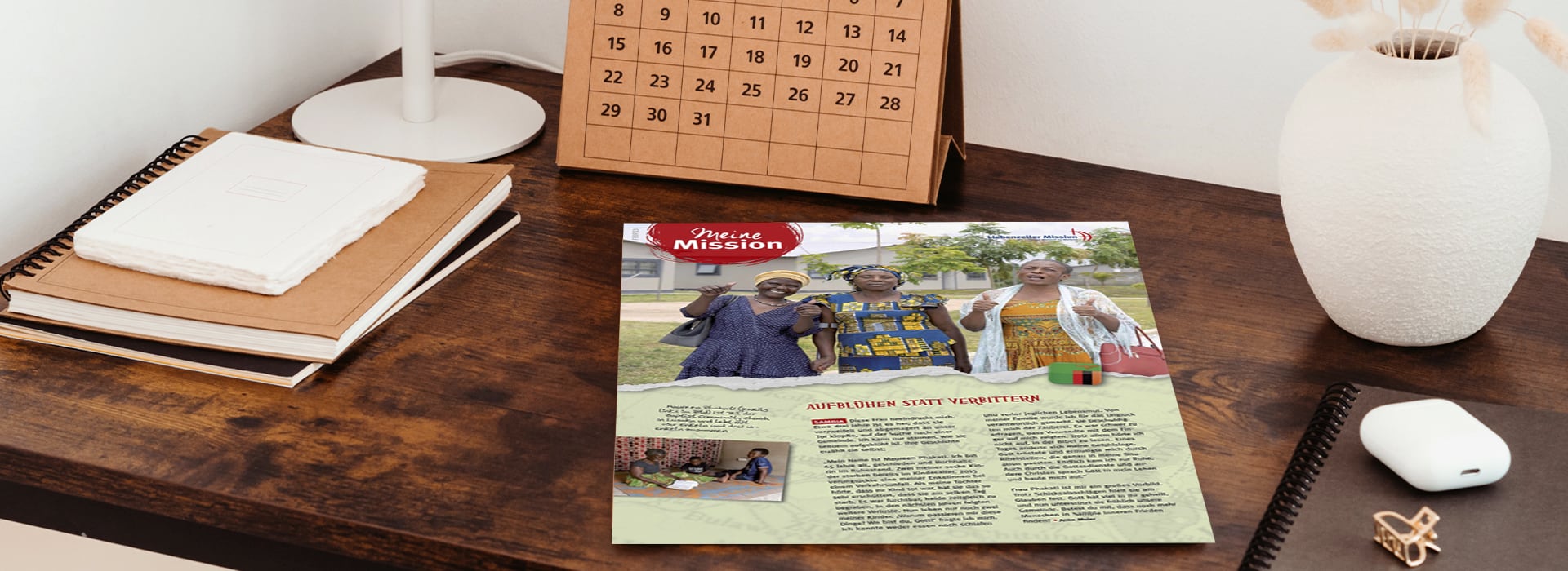 Auf einem Holzschreibtisch, auf dem Notizbücher, ein Kalender und eine weiße Vase liegen, steht ein Newsletter, auf dem drei Frauen abgebildet sind.