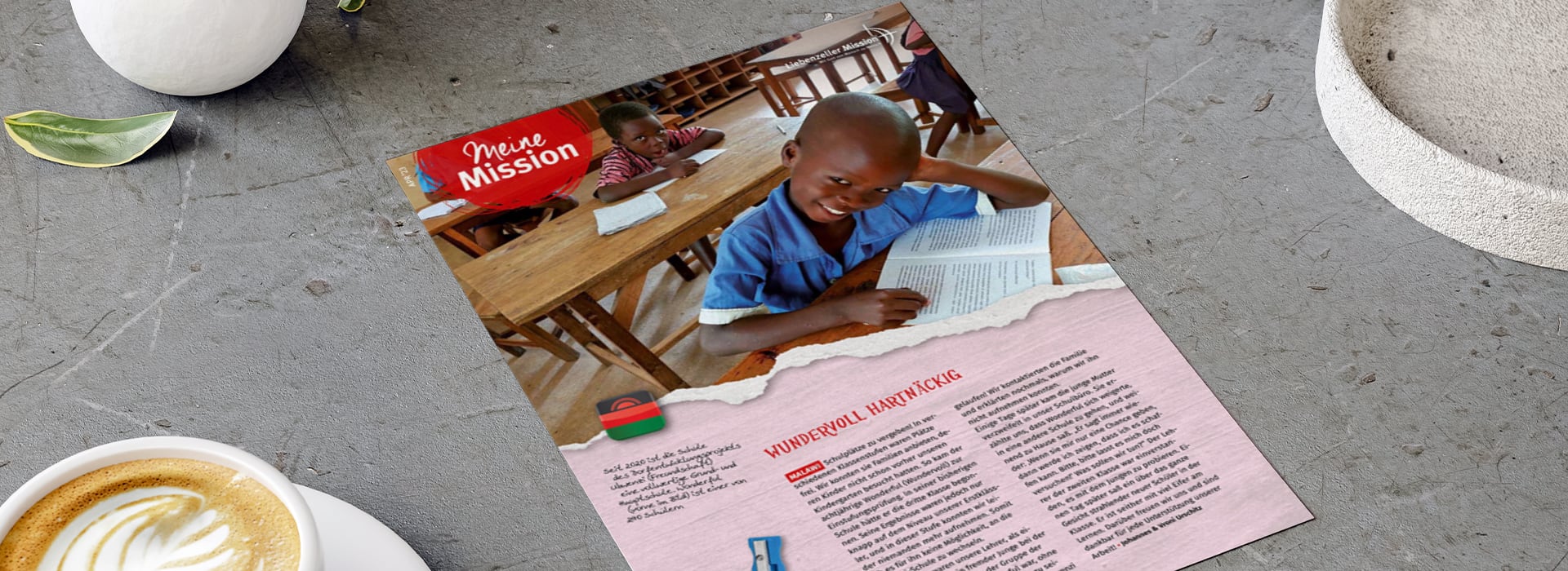 Eine Zeitschriftenseite, auf der ein lächelndes Kind in einem Klassenzimmer zu sehen ist, das an einem Holztisch ein Buch liest; daneben stehen Kaffee und eine Pflanze.