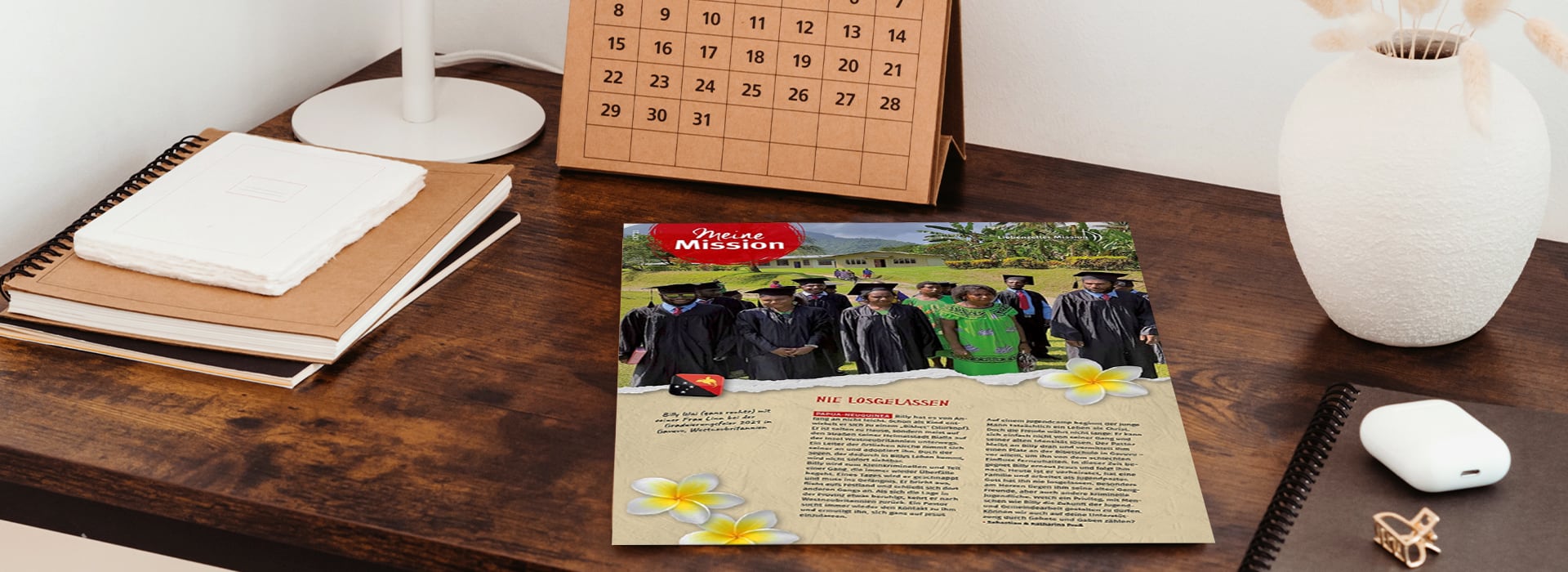 Ein Magazin mit einem Gruppenfoto vom Schulabschluss auf einem Holztisch neben einem Kalender, Notizbüchern und einer weißen Vase.