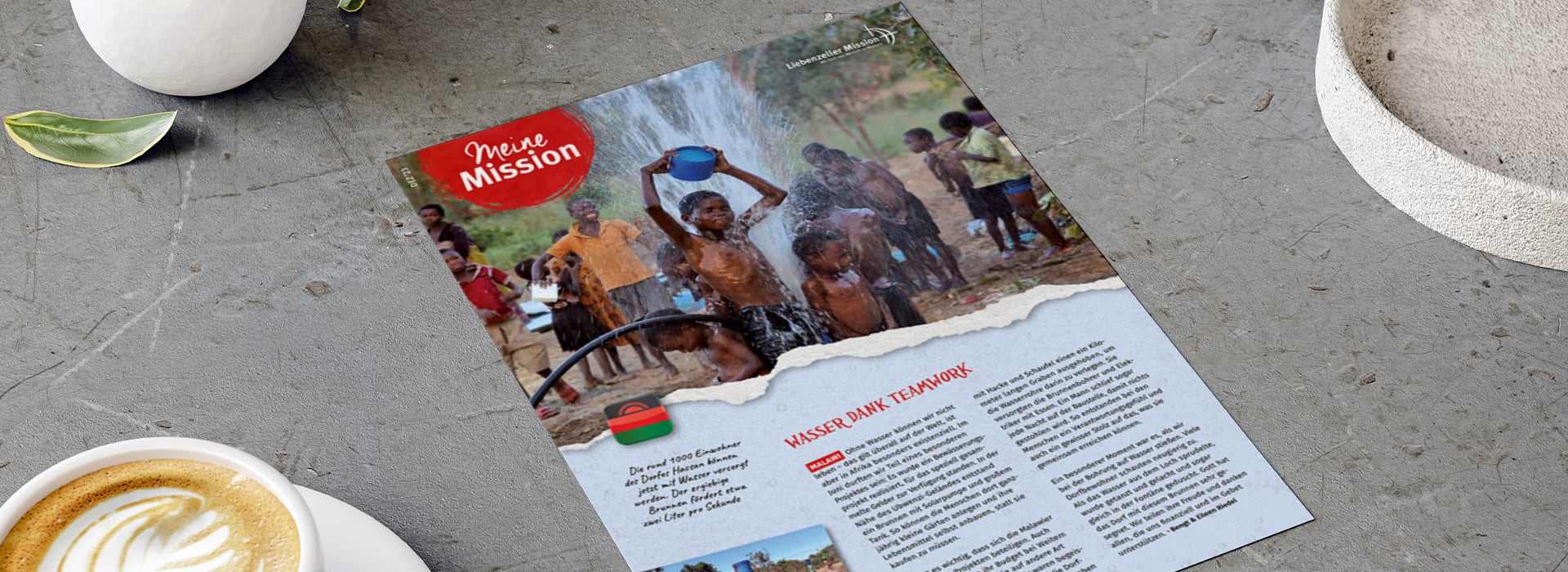 Auf einer Zeitschriftenseite, die auf einem Tisch liegt, sind Kinder zu sehen, die im Wasser spielen; daneben stehen eine Kaffeetasse und eine Pflanze.