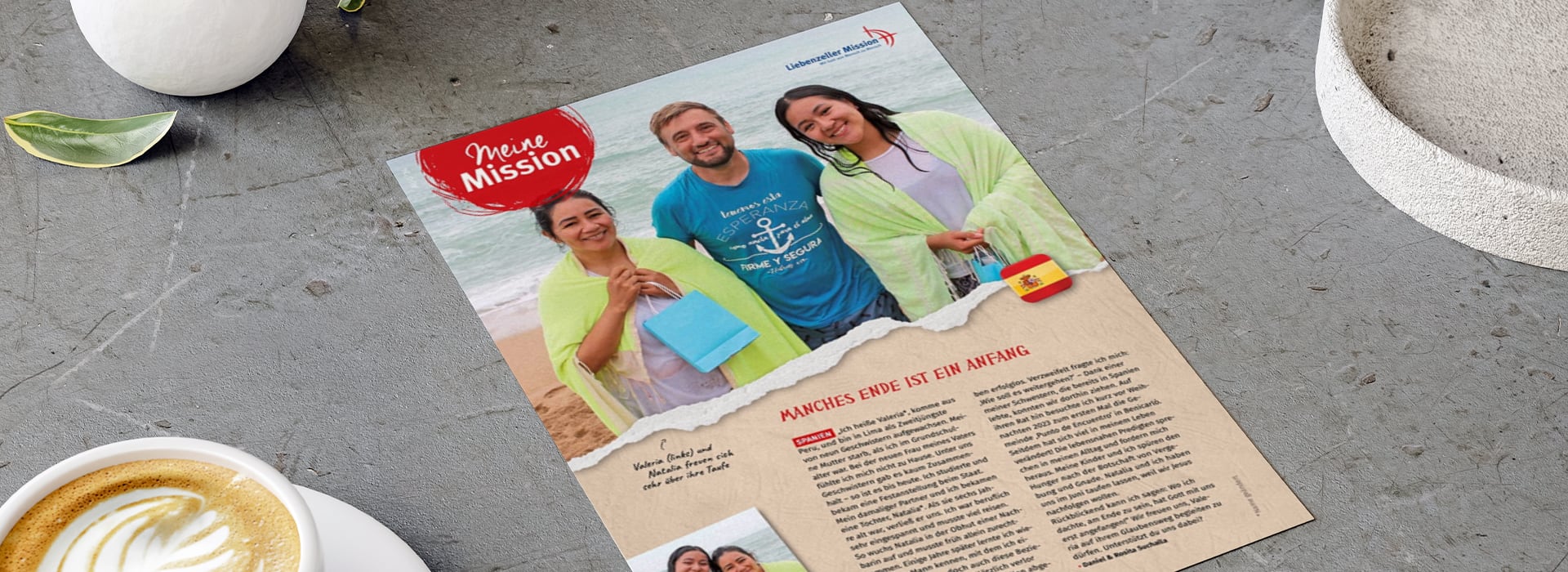 Auf einer Zeitschriftenseite, die auf einem grauen Tisch liegt, sind drei lächelnde Menschen zu sehen, die Geschenke in den Händen halten; unten sind ein Text und eine kleine spanische Flagge abgebildet.