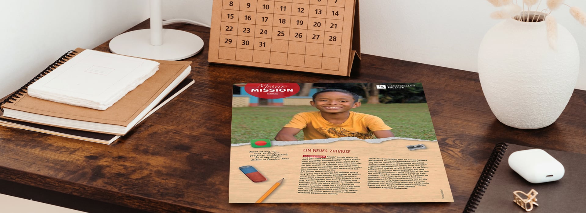 Auf einem Holztisch liegen eine Zeitschriftenseite mit einem lächelnden Jungen, Bleistifte und ein Radiergummi neben einem Kalender, einem Notizbuch und einer Vase.
