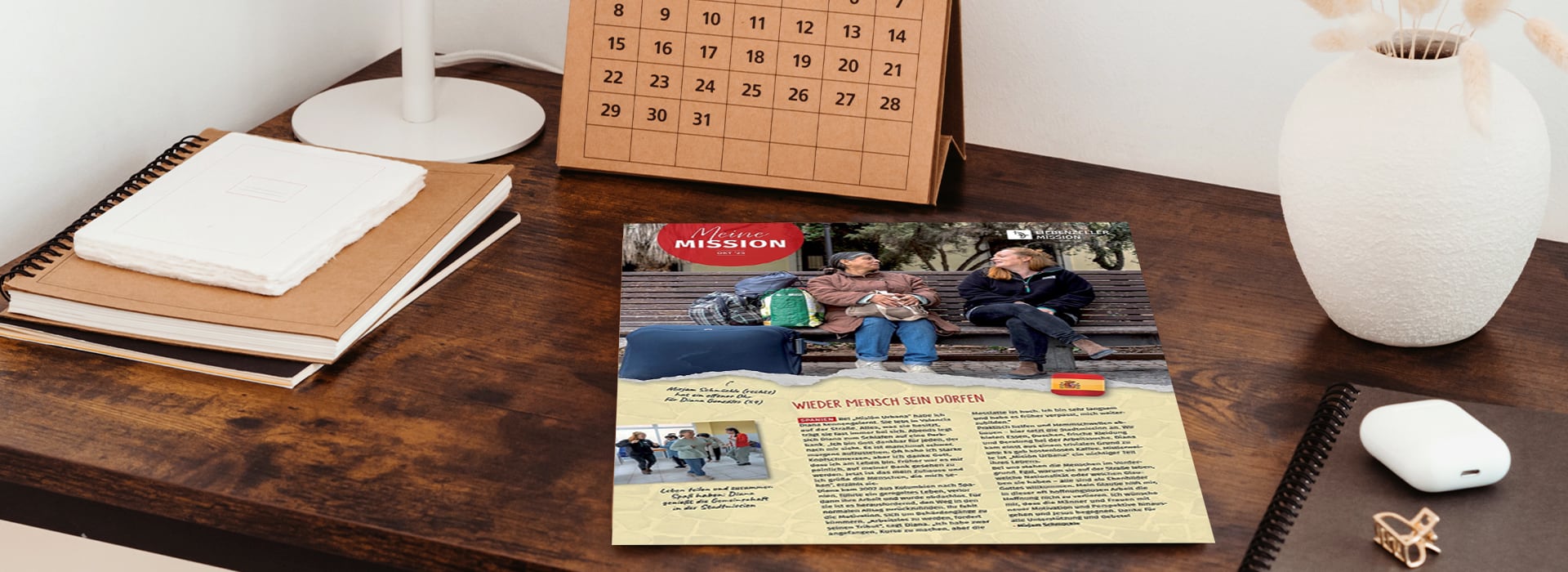 Auf einem Holzschreibtisch liegt eine Zeitschrift aufgeschlagen, daneben stehen eine Lampe, ein Kalender und Notizbücher.