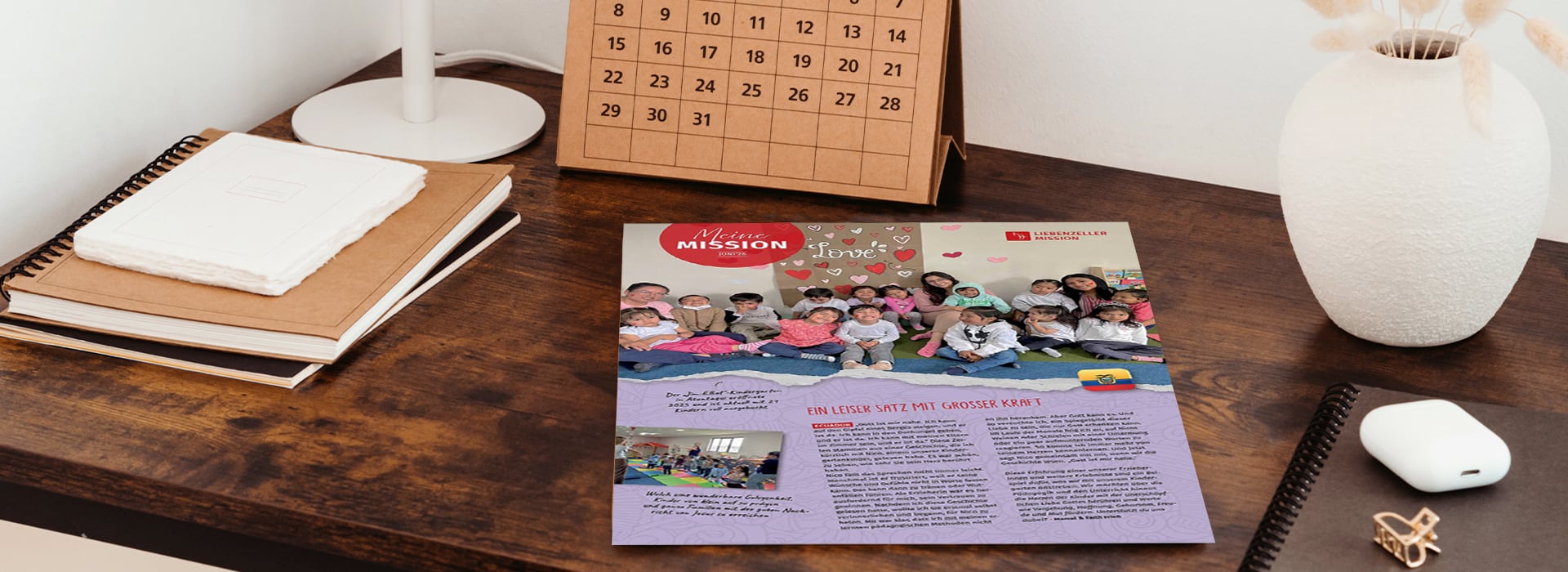 Auf einem Holztisch liegt eine Zeitschrift mit einem Gruppenfoto von Kindern neben einem Kalender, einem Notizbuch und einer Topfpflanze.