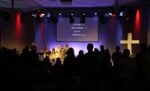 Ein Gottesdienst mit Menschen, die auf der Bühne singen; auf einem Bildschirm über ihnen wird "Danke" in mehreren Sprachen angezeigt.