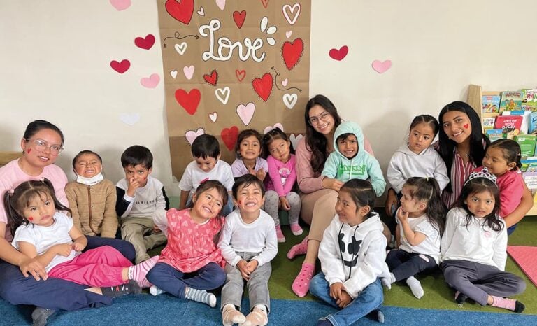 Eine Gruppe kleiner Kinder und Erwachsener sitzt lächelnd vor einer Wand, die mit Herzen und dem Wort „Love“ verziert ist.