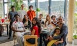 Eine bunte Gruppe von Menschen, Erwachsene und Kinder, sitzt und steht lächelnd in einem hellen Café mit großen Fenstern.