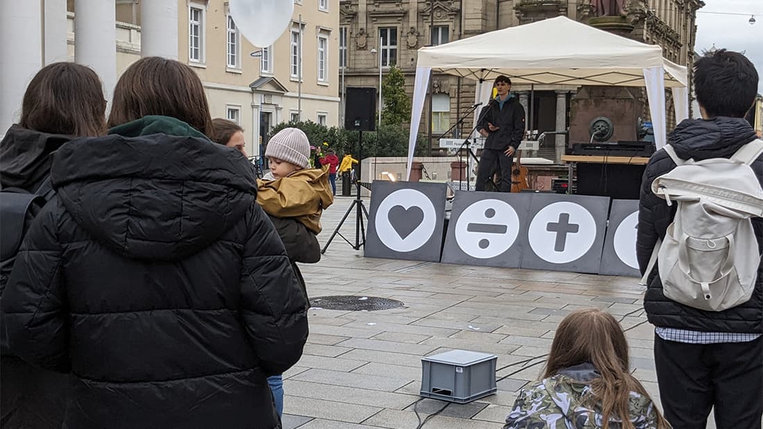 Ein Mann hält auf einer Freiluftbühne mit Mathematik- und Herzsymbolen eine Rede, während die Menschen auf einem Stadtplatz zuschauen.