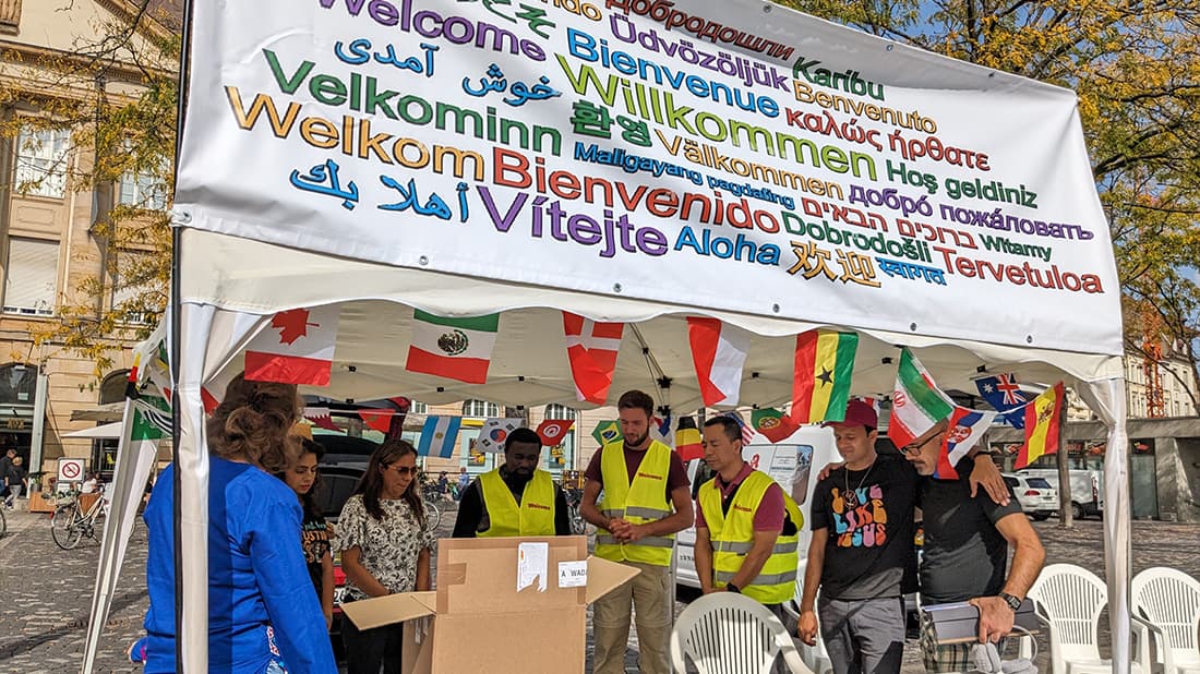 Menschen in gelben Westen stehen unter einem Zelt, über dem in vielen Sprachen „Willkommen“ steht und Fahnen wehen.