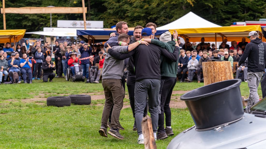 Eine Gruppe von Männern umarmt sich und feiert im Freien, während eine Menschenmenge bei einer Veranstaltung mit Zelten und verstreuten Reifen zusieht.