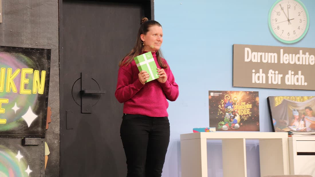 Eine Frau in einem rosa Pullover hält eine grüne Geschenkbox auf einer Bühne in der Hand, im Hintergrund sind Bücher und eine Uhr zu sehen.