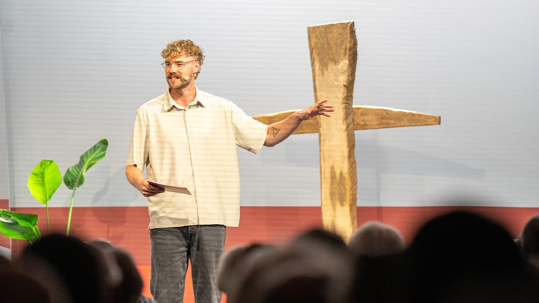 Ein Mann mit Brille spricht auf der Bühne und deutet auf ein großes Holzkreuz; im Vordergrund sind eine Pflanze und das Publikum zu sehen.