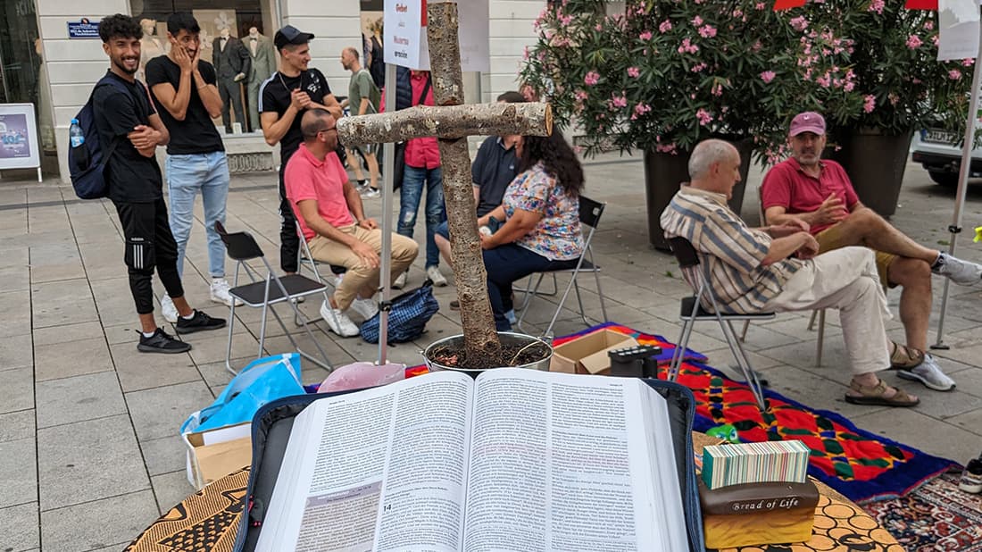 Ein offenes Buch auf einem Tisch im Freien, daneben sitzen Menschen in Gruppen und unterhalten sich, und ein Baumast ist quer darüber gelegt.