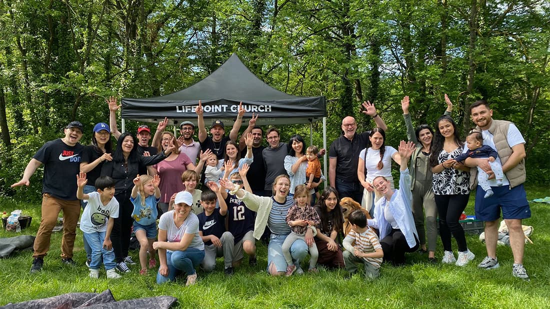 Eine bunt gemischte Gruppe von Menschen lächelt und winkt, während sie sich im Freien vor einem Vordach der „Life Point Church“ versammelt hat.