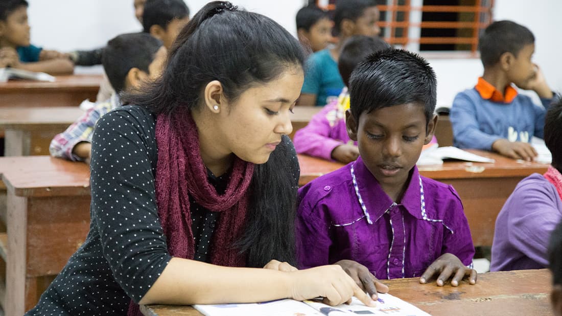 Ein Lehrer hilft einem Schüler beim Lesen eines Buches in einem Klassenzimmer, während im Hintergrund andere Kinder an ihren Schreibtischen sitzen.