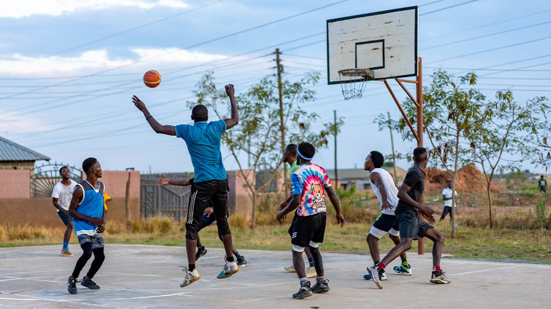 Eine Gruppe von Männern spielt draußen Basketball; einer springt hoch, um den Ball zu blocken, während die anderen zuschauen und sich darauf vorbereiten, zu reagieren.