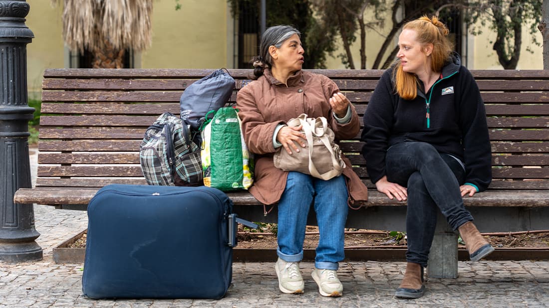 Zwei Frauen sitzen auf einer Bank und unterhalten sich, umgeben von Taschen und einem Koffer, im Freien in einer städtischen Umgebung.