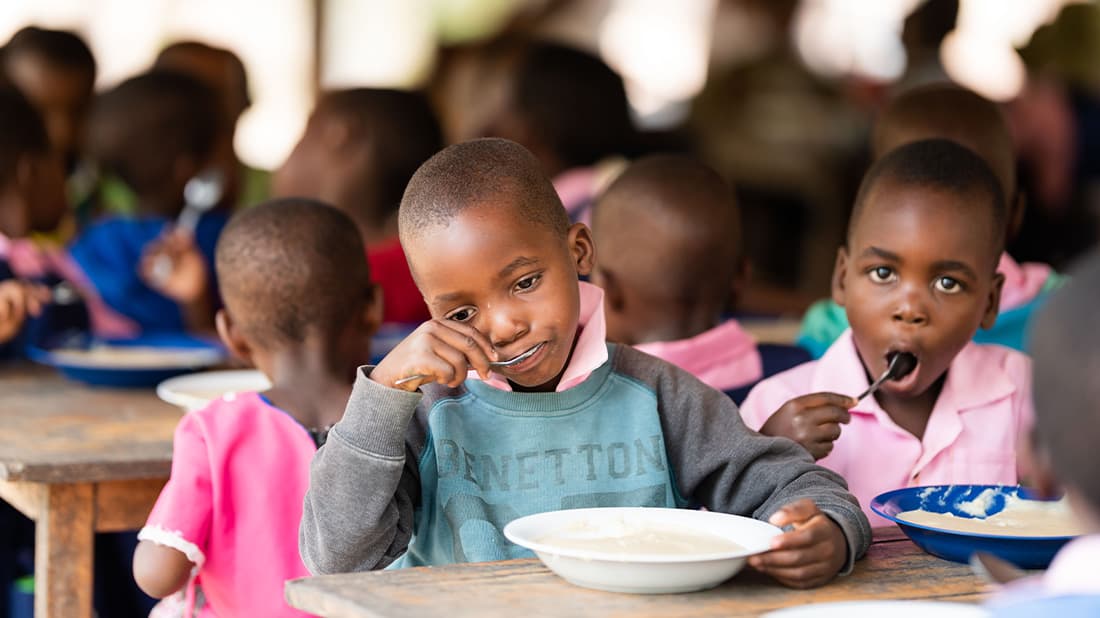 Kleine Kinder sitzen an einem Tisch und essen aus Schüsseln; ein Junge lächelt in die Kamera.