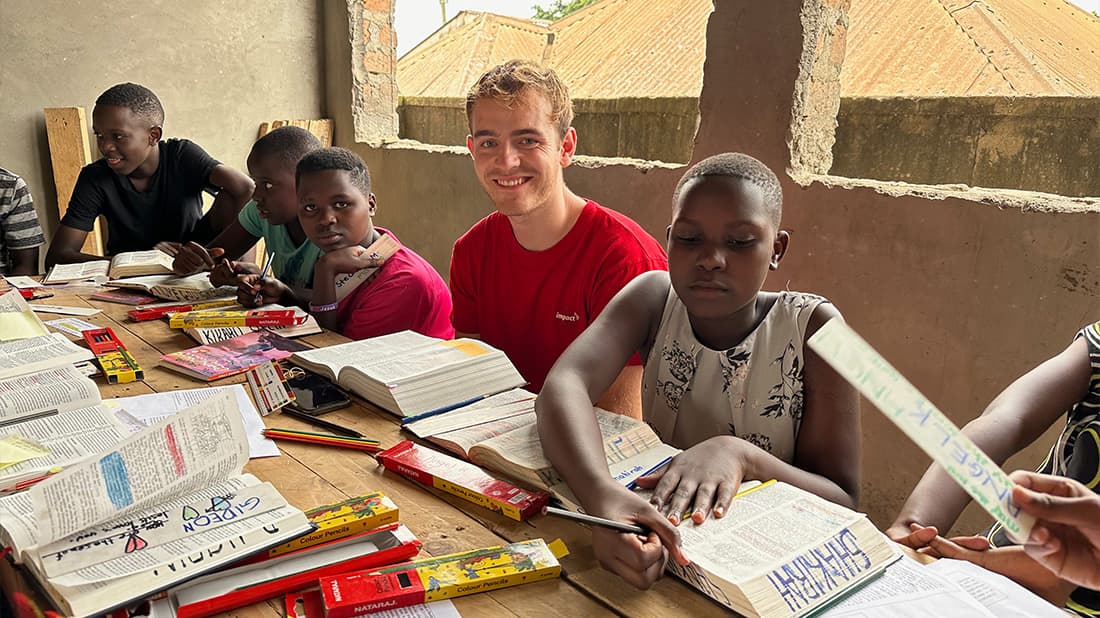 Eine Gruppe von Kindern und ein junger Mann sitzen an einem Tisch und lesen und schreiben mit aufgeschlagenen Büchern und Schulmaterial.
