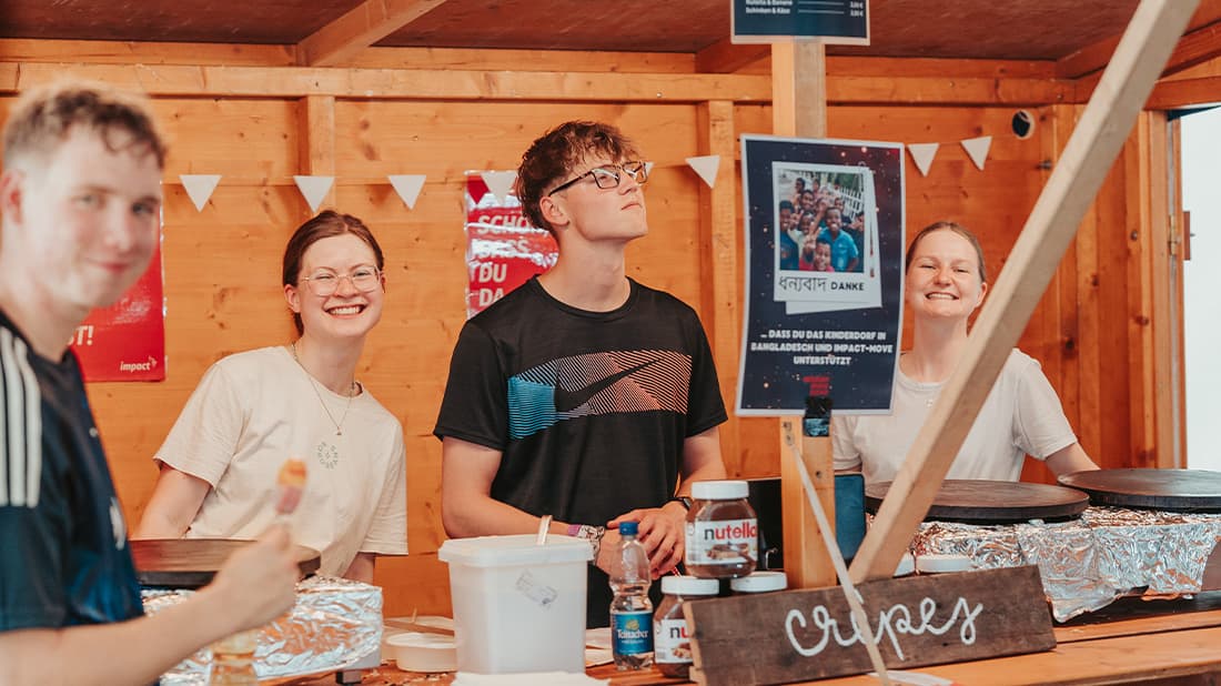 Hinter einem Imbissstand mit Crêpes-Pfannen, Nutella-Gläsern und einem Holzschild mit der Aufschrift „Crêpes“ stehen vier lächelnde junge Leute.