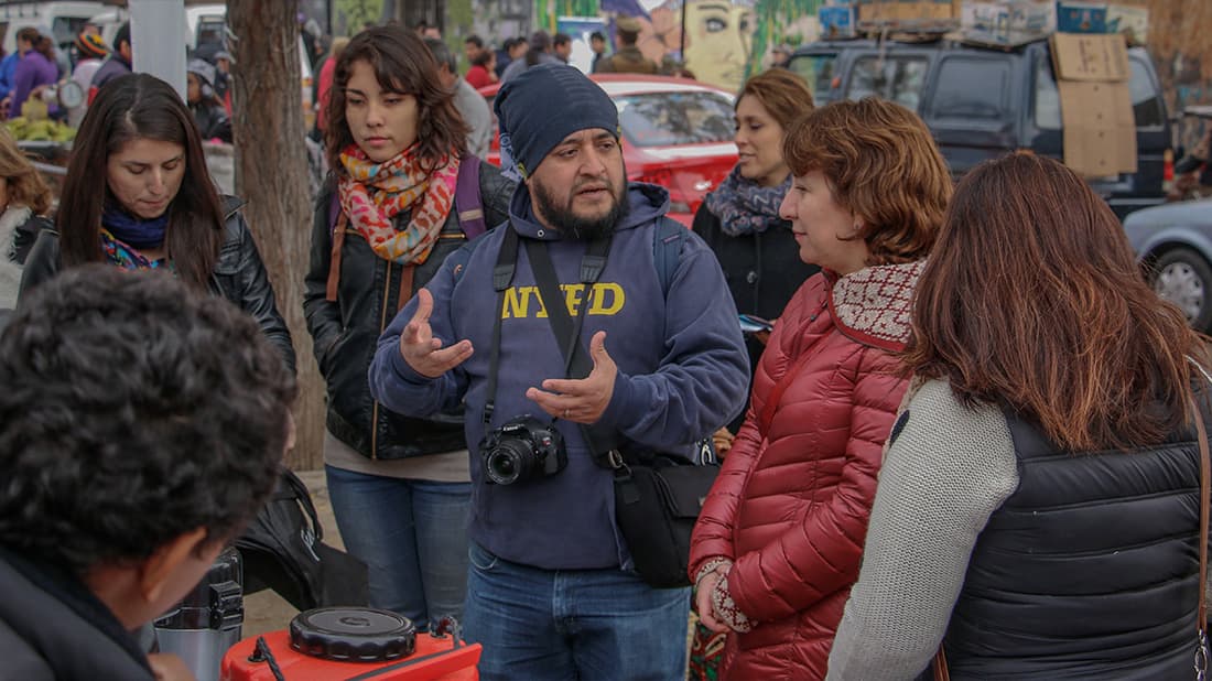 Eine Gruppe von Menschen unterhält sich im Freien; ein Mann trägt eine Kamera und einen „Nafed“-Hoodie, die anderen hören aufmerksam zu.