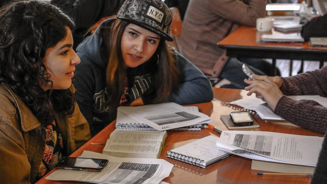 Zwei junge Frauen unterhalten sich an einem Tisch, der mit Notizbüchern, Papieren und Handys bedeckt ist; im Hintergrund arbeiten andere.