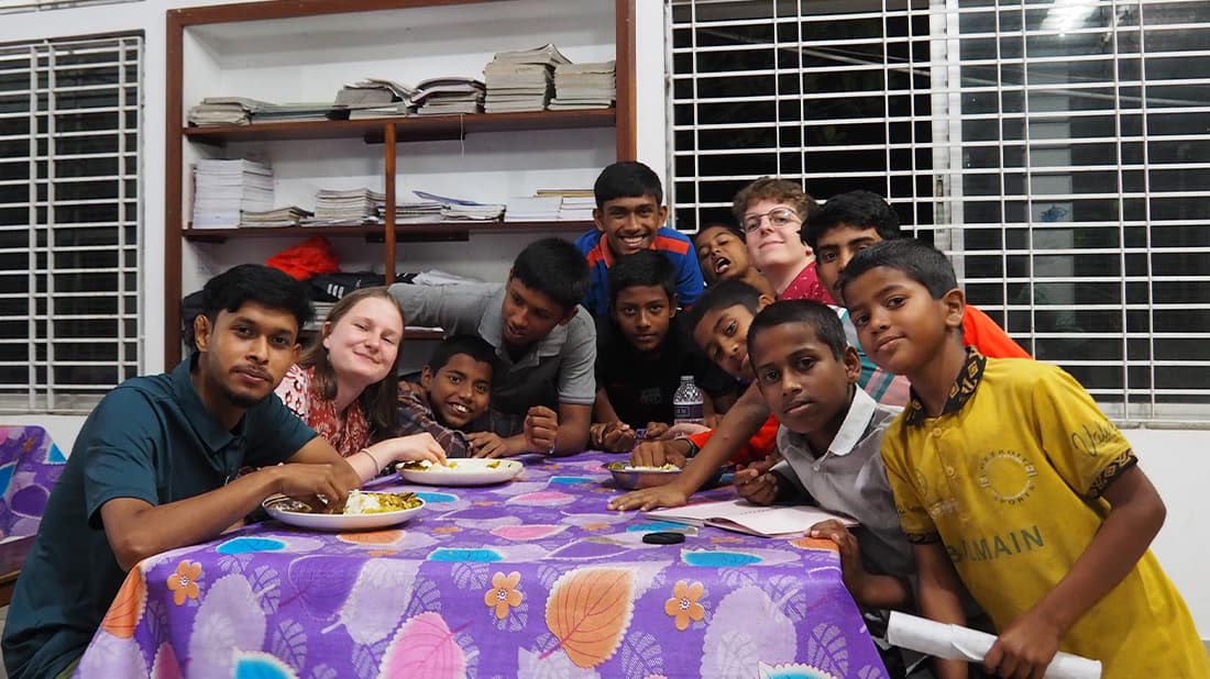 Eine Gruppe von Kindern und zwei Erwachsene sitzen lächelnd um einen Tisch mit Essen herum, in einem Raum mit Regalen und vergitterten Fenstern.