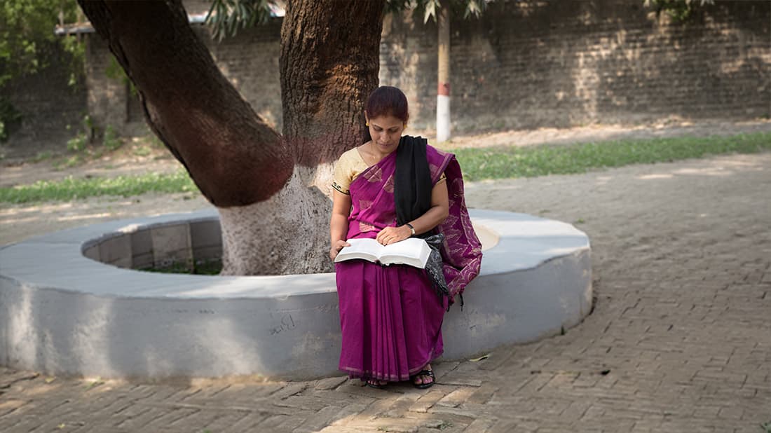 Eine Frau in einem violetten Sari sitzt auf einer runden Bank um einen Baum herum und liest im Freien ein Buch.