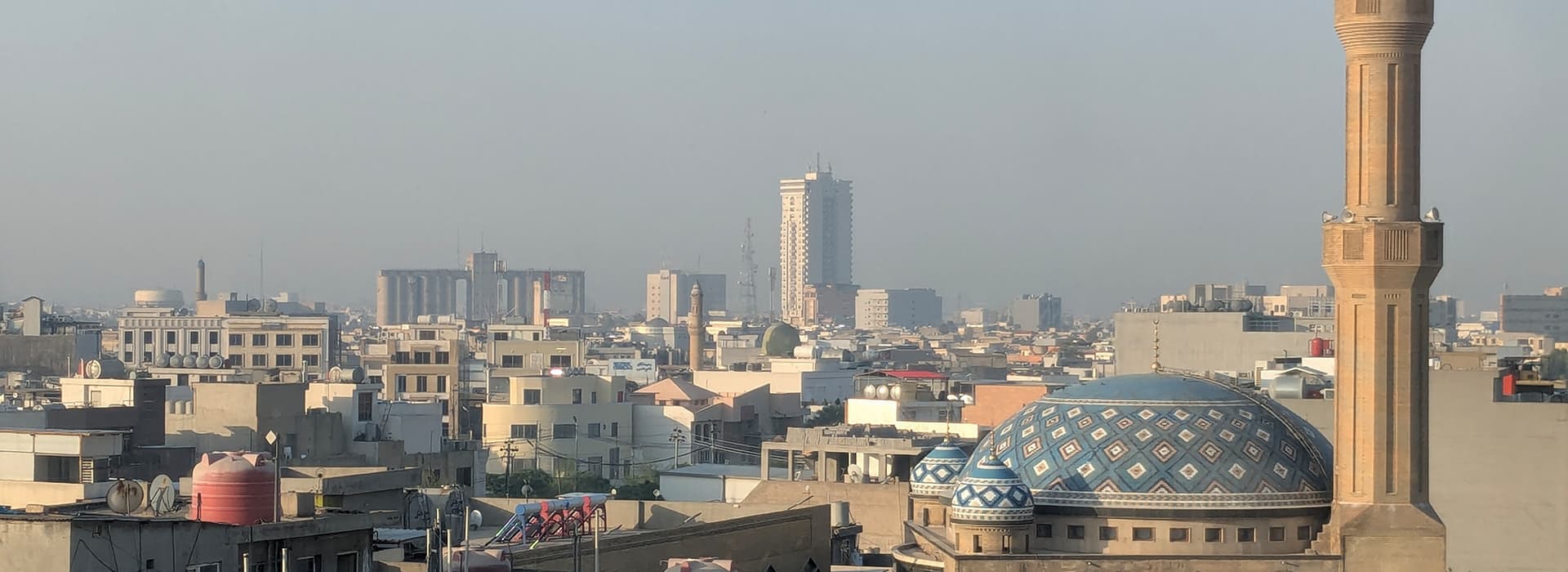 Stadtansicht mit vielen Gebäuden, einer Moschee mit blauer Kuppel und einem hohen Minarett unter einem dunstigen Himmel.