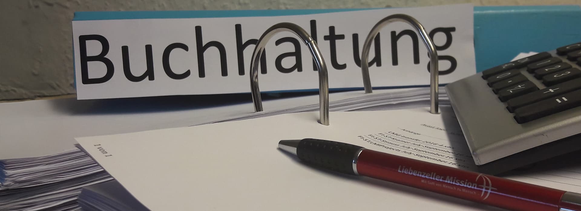 Ein Ordner mit der Aufschrift „Buchhaltung“, Papiere, ein roter Stift und ein Taschenrechner auf einem Schreibtisch.