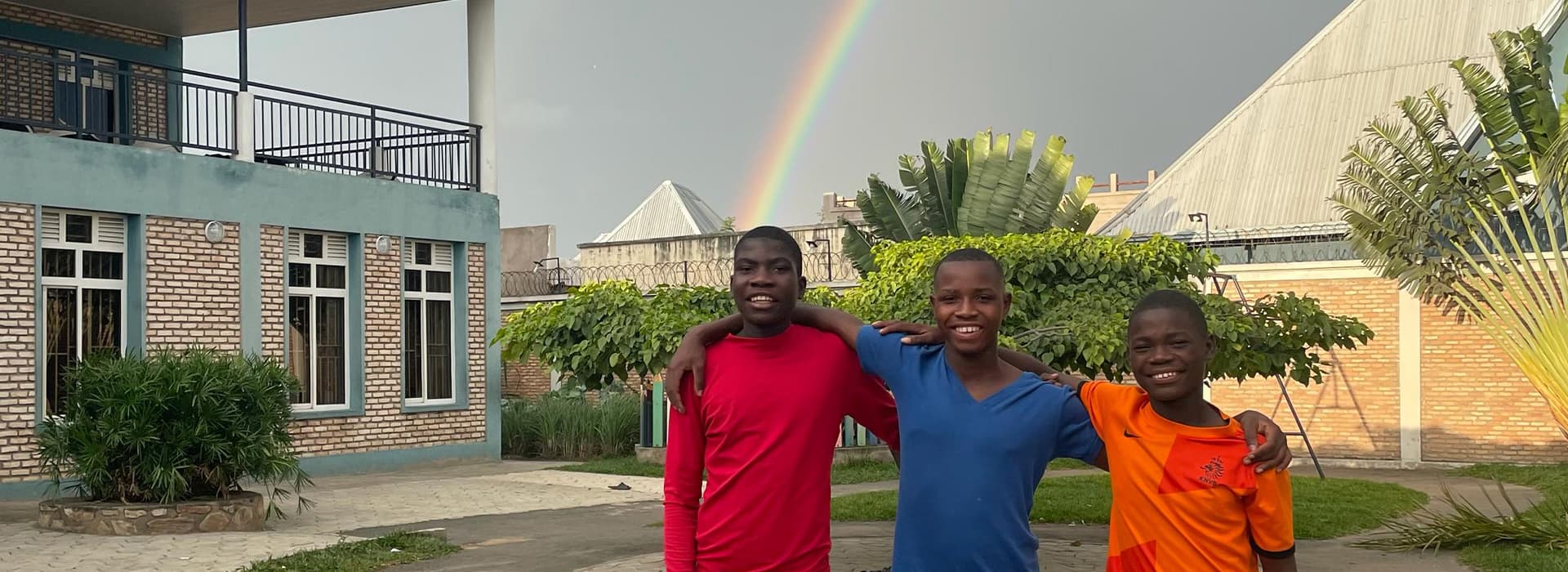Drei Jungen stehen mit umeinander gelegten Armen vor einem Gebäude, hinter ihnen ist ein Regenbogen am Himmel zu sehen.