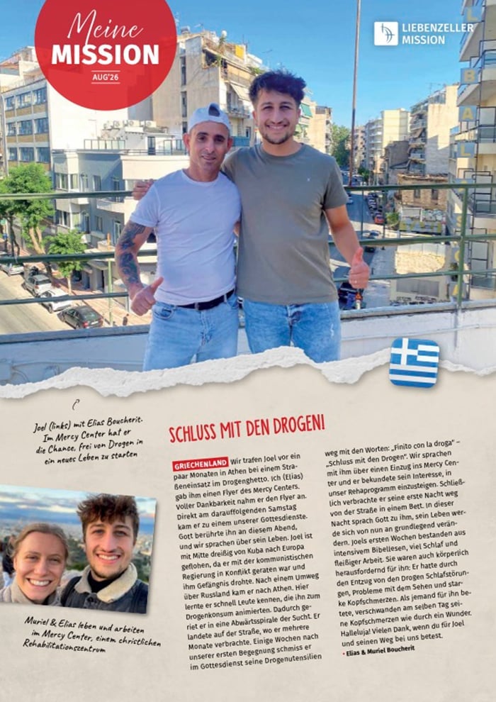 Zwei Männer stehen lächelnd nebeneinander auf einem Balkon, im Hintergrund sind Gebäude zu sehen und im Vordergrund weht eine griechische Flagge.
