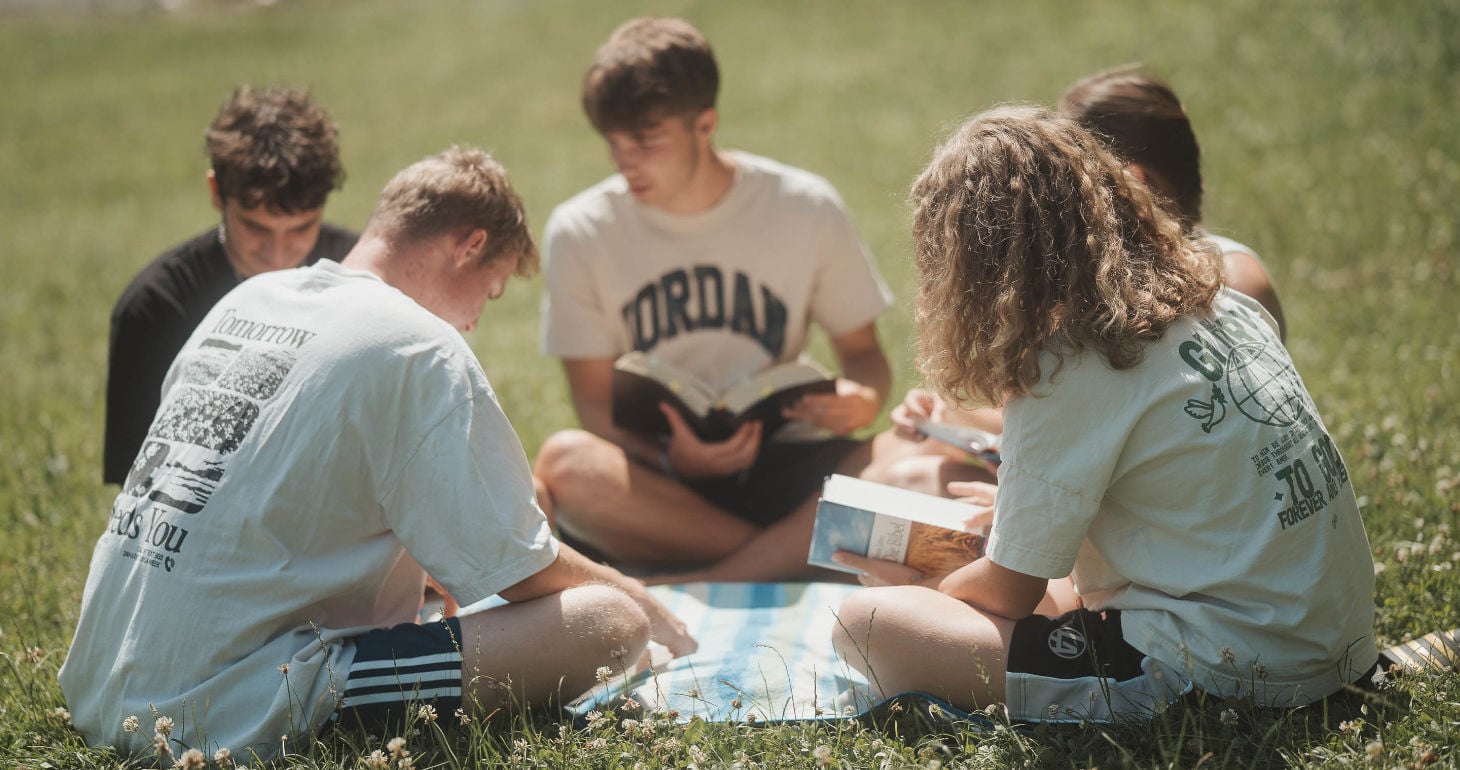 Fünf Menschen sitzen auf dem Rasen und lesen gemeinsam auf einer gestreiften Decke im Sonnenlicht Bücher.