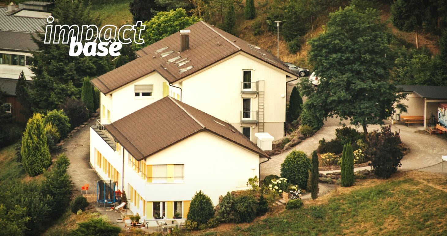Ein großes, cremefarbenes Haus mit braunem Dach, umgeben von Bäumen und Sträuchern, mit überlagertem Basistext in Fettdruck.