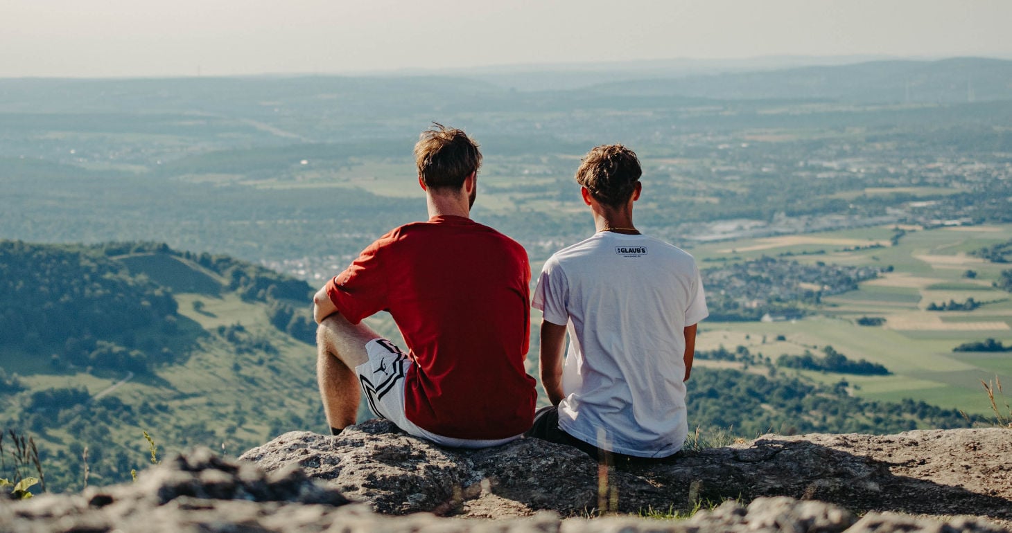 An einem klaren Tag sitzen zwei Menschen auf einem Felsvorsprung und blicken auf eine malerische Landschaft aus Feldern und Hügeln.