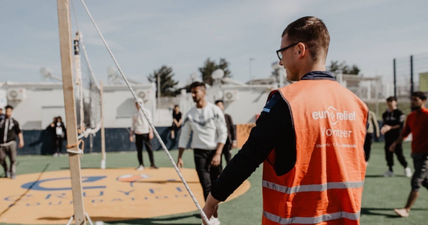 Ein Freiwilliger in einer orangefarbenen Weste beobachtet, wie Menschen auf einem Außenplatz Volleyball spielen.