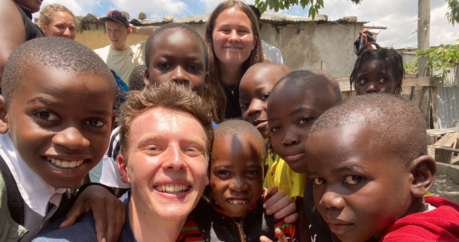 Eine Gruppe lächelnder Kinder und zwei Erwachsene posieren an einem sonnigen Tag im Freien dicht beieinander für ein Selfie.