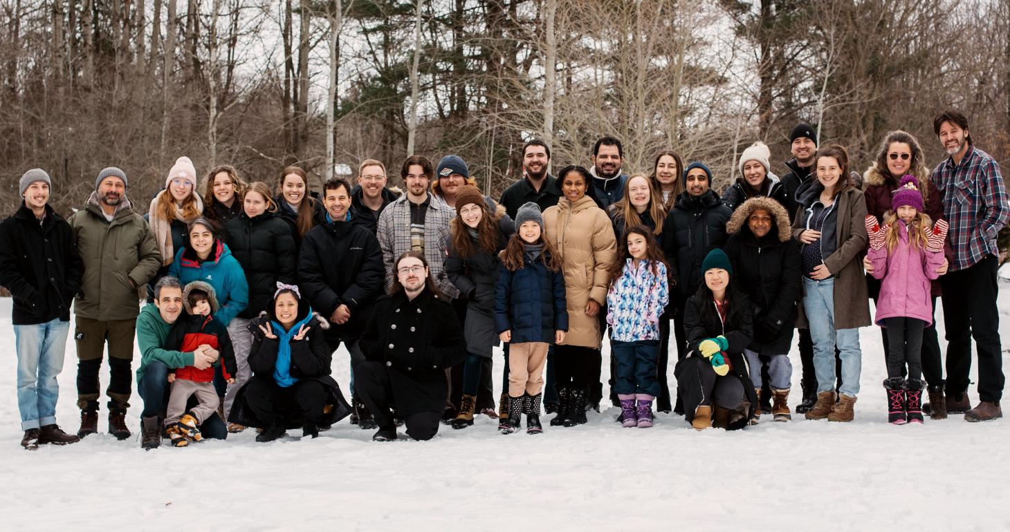Eine große Gruppe von Menschen posiert gemeinsam im Freien in Winterkleidung, sie stehen im Schnee, im Hintergrund sind Bäume zu sehen.