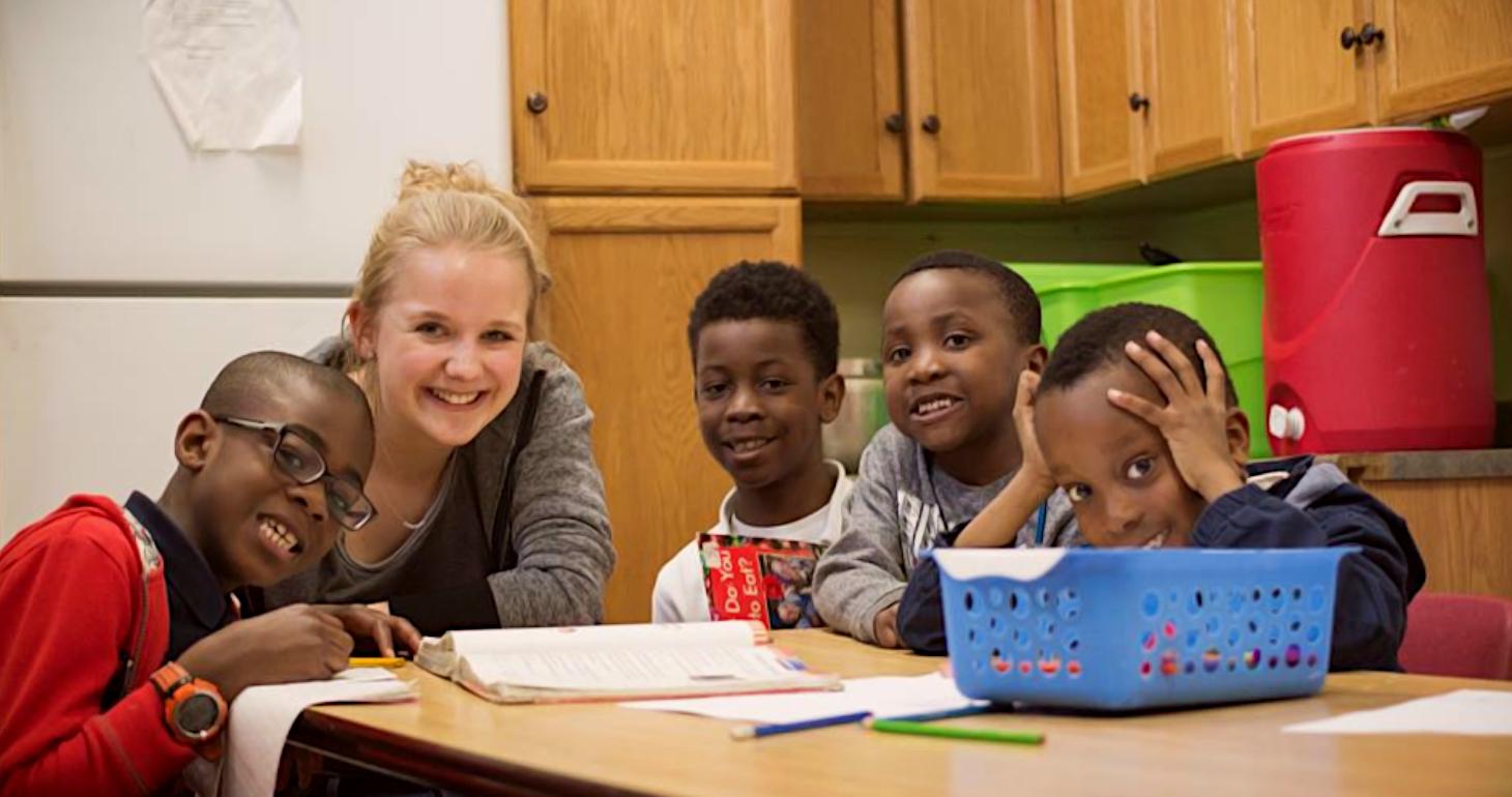 Fünf lächelnde Kinder und ein Erwachsener sitzen an einem Tisch mit Papier und Stiften in einem Klassenzimmer oder einer Küche.