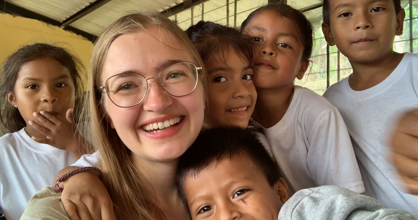 Eine lächelnde junge Frau mit Brille, umgeben von fünf Kindern in einem hellen Raum; alle blicken in die Kamera.