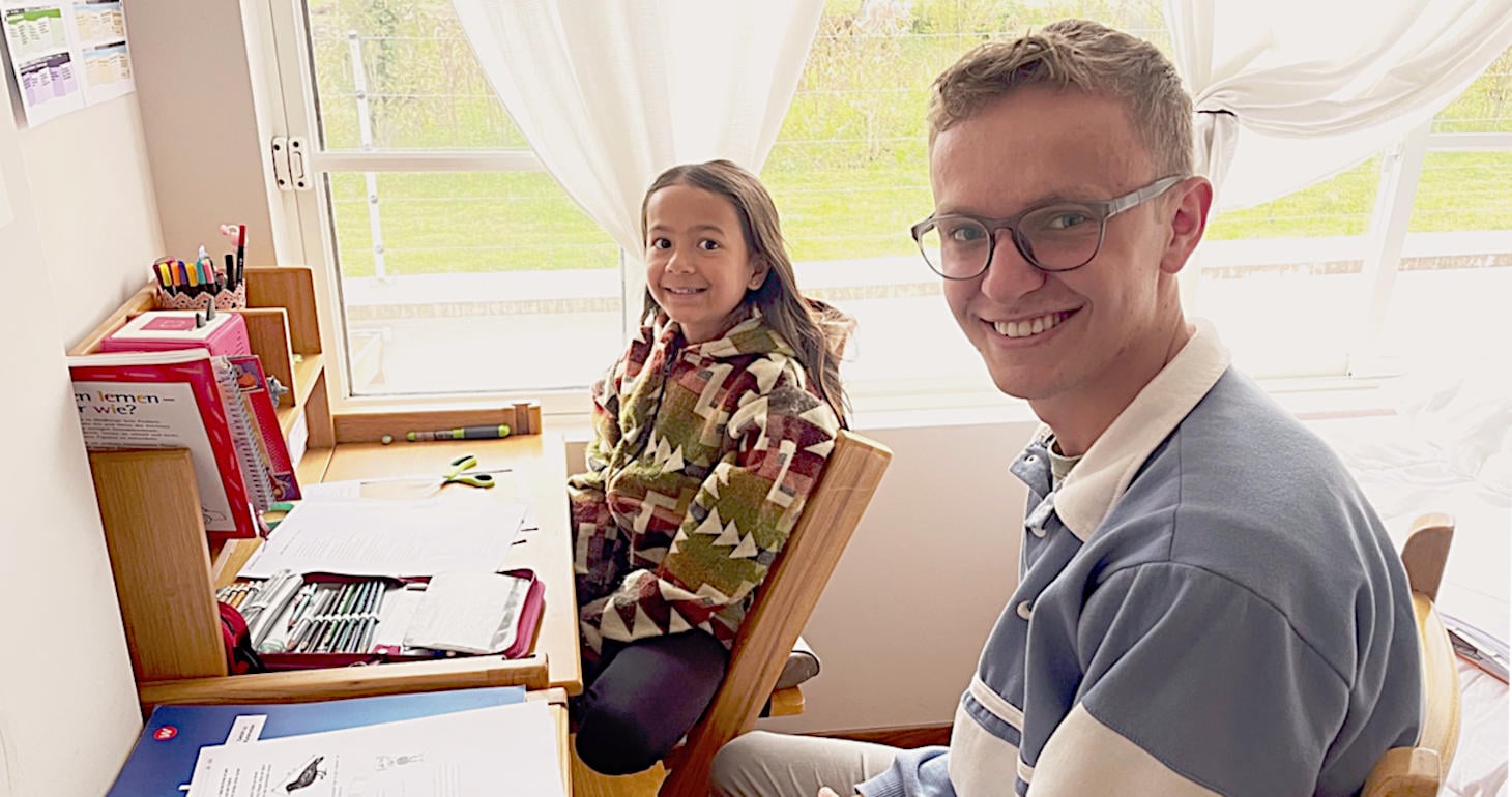 Ein lächelnder Mann und ein Mädchen sitzen an einem Schreibtisch mit Papieren und Schulmaterial in der Nähe eines hellen Fensters.