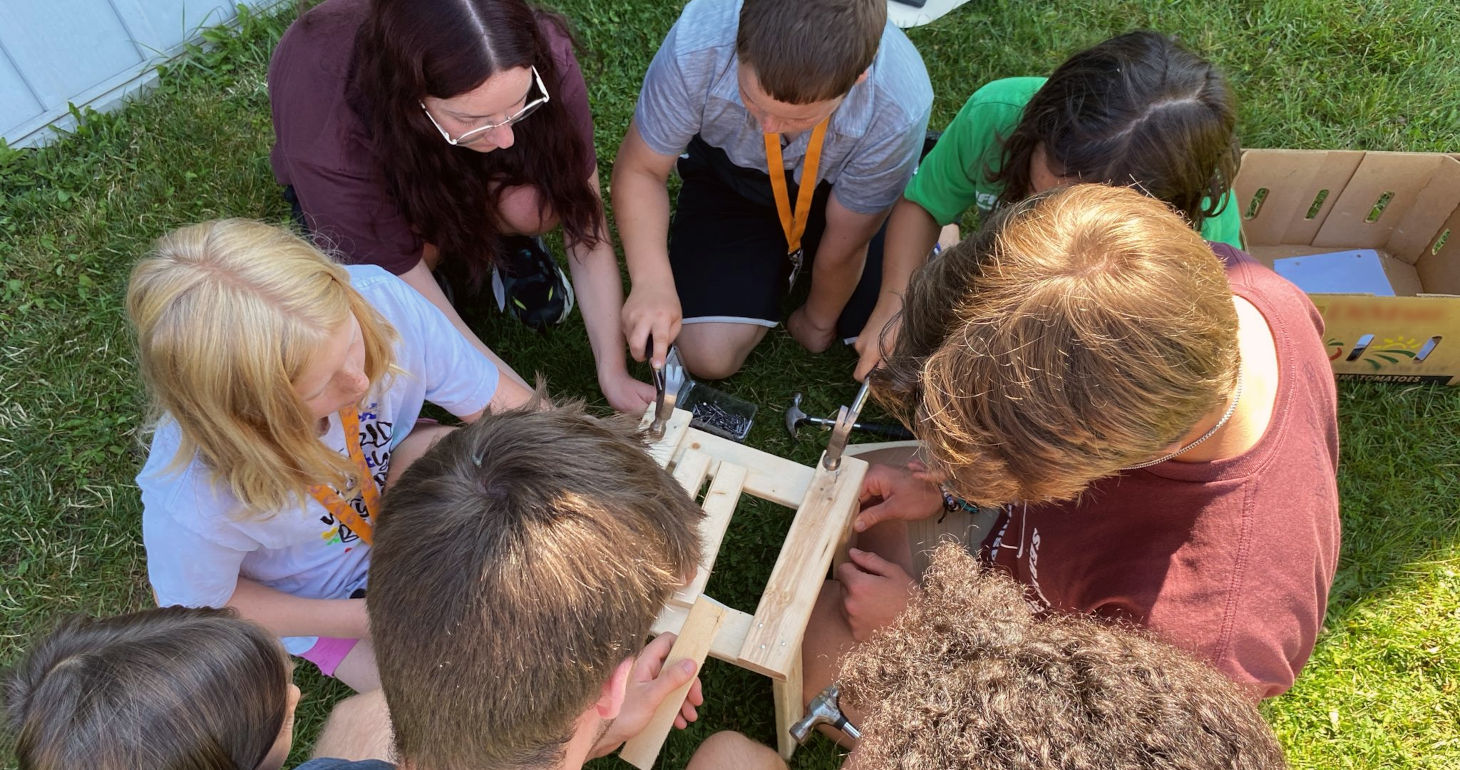 Sieben Kinder bauen auf einer Wiese eine kleine Holzkonstruktion auf, arbeiten dabei zusammen und blicken auf ihr Projekt hinunter.