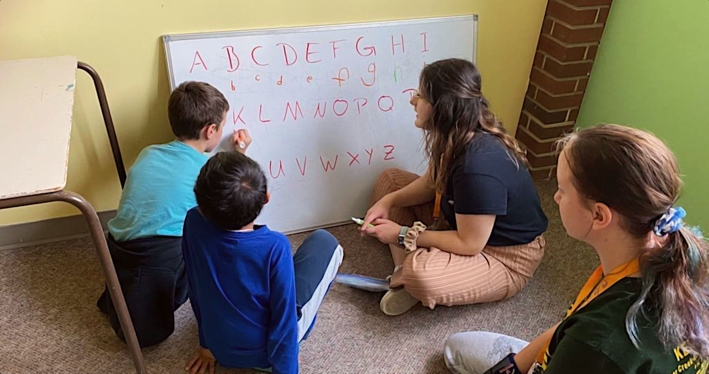 Drei Kinder und ein Erwachsener sitzen vor einer Weißwandtafel und üben gemeinsam das Schreiben von Groß- und Kleinbuchstaben.