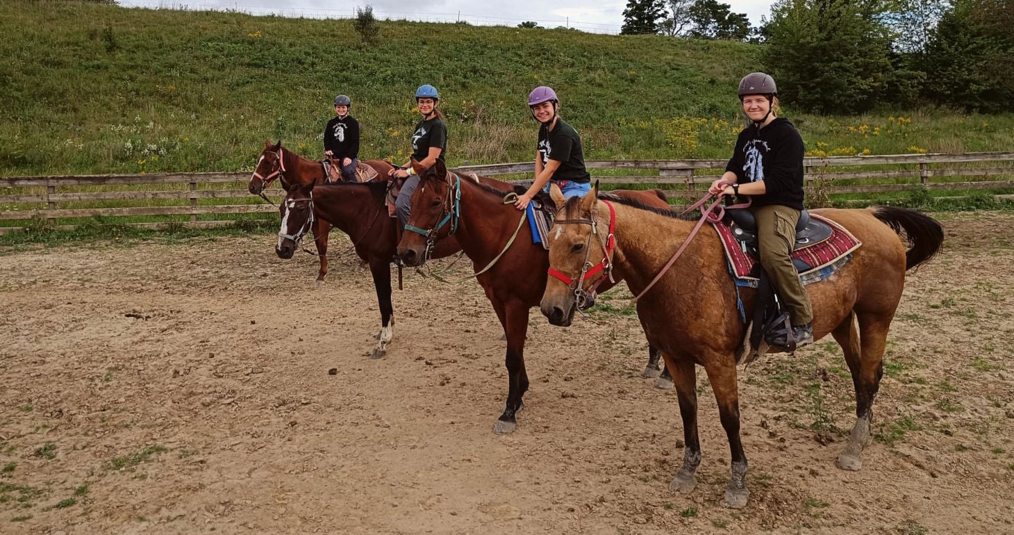 Vier Personen mit Helmen und schwarzen Hemden sitzen auf Pferden in einem eingezäunten Reitplatz im Freien, hinter ihnen liegen grasbewachsene Hügel.
