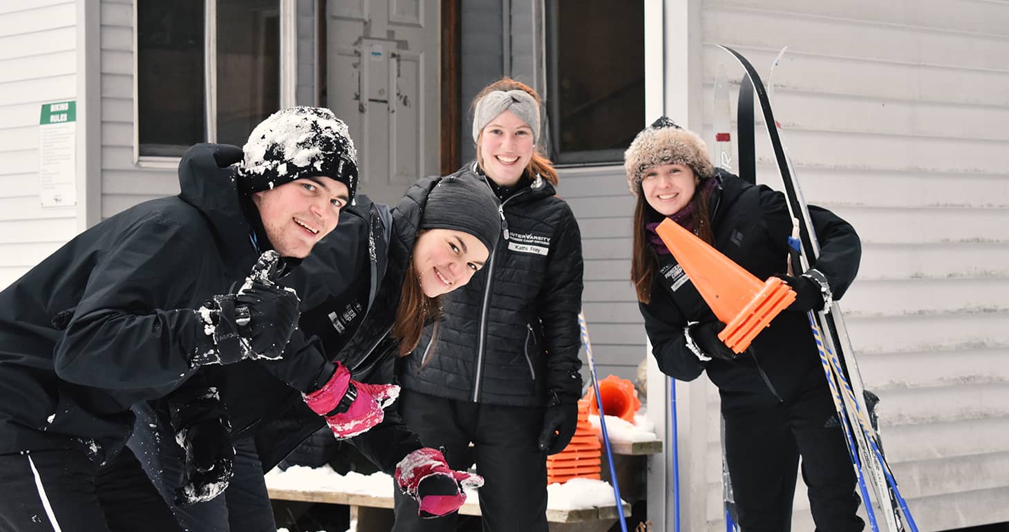 Vier Personen in Winterkleidung lächeln im Freien, halten Skistöcke und orangefarbene Kegel in den Händen, und auf ihren Mützen und Jacken liegt Schnee.