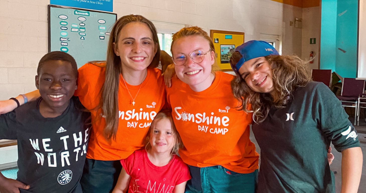 Fünf lächelnde Kinder und Jugendliche, von denen zwei orangefarbene „Sunshine Day Camp“-T-Shirts tragen, posieren gemeinsam in einem Raum und haben ihre Arme umeinander gelegt.