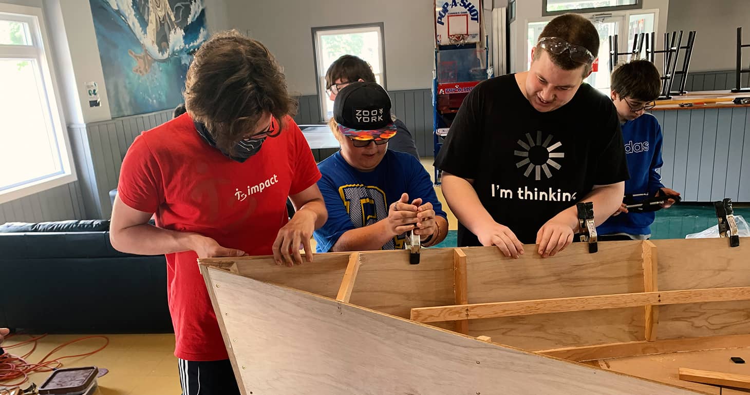 Drei Jugendliche bauen in einem Raum mit Hilfe von Klemmen und Werkzeugen eine Holzkonstruktion zusammen und sind ganz auf ihr praktisches Projekt konzentriert.